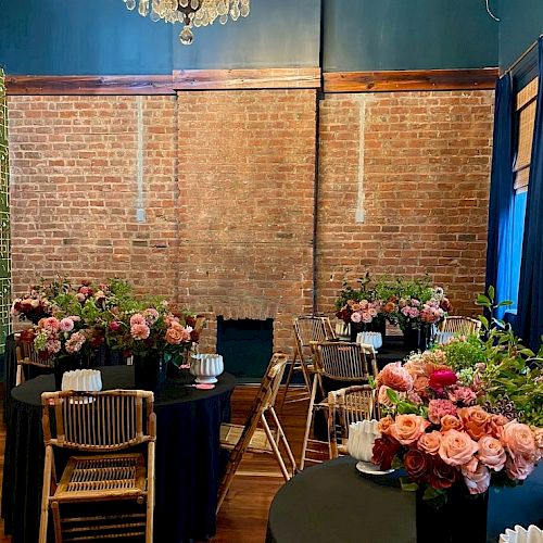 A brick-walled room with tables, flowers, and chairs, featuring a chandelier hanging above, creating an elegant atmosphere.