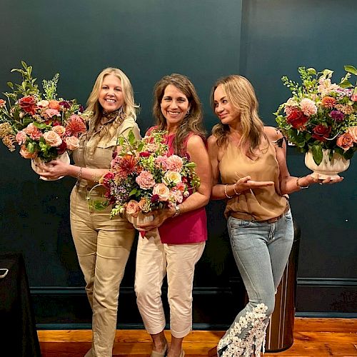 Three people smiling, holding large colorful flower arrangements, standing in front of a dark wall.
