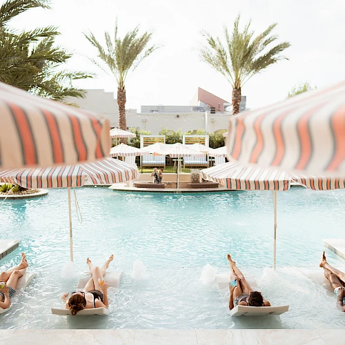People lounging on pool loungers under striped umbrellas around a sunny pool, palm trees in the background.