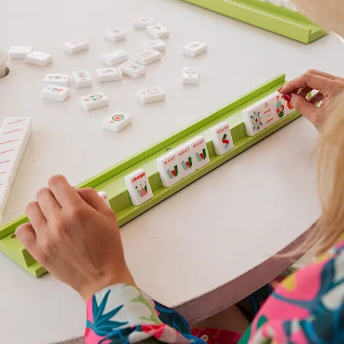 A person’s hands are placing small game tiles/cards along a green board in a tabletop board game, with other tiles scattered around.