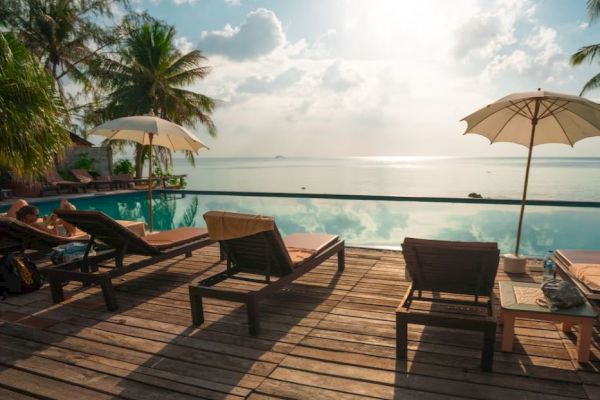 Beachfront area with lounge chairs, umbrellas, and a pool overlooking the ocean under a partly cloudy sky.