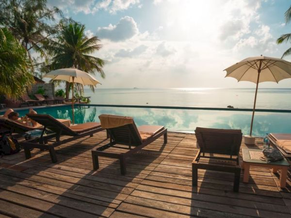 Beachfront area with lounge chairs, umbrellas, and a pool overlooking the ocean under a partly cloudy sky.