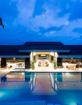 A luxurious building with open-air seating areas overlooks a large swimming pool, all illuminated under a vibrant evening sky.