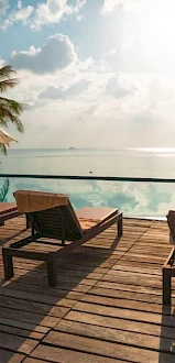 Poolside chairs and umbrellas on a wooden deck, overlooking a calm sea with palm trees and sunlit clouds in the background.