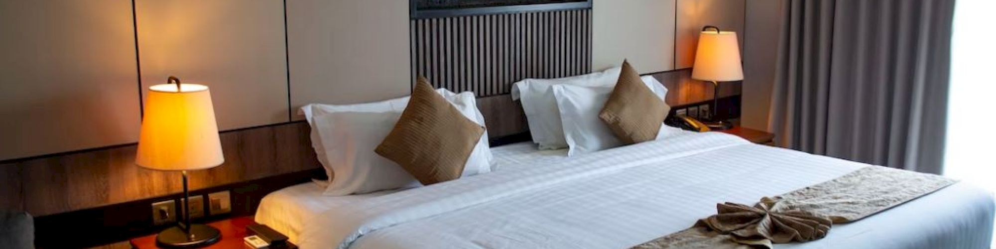 A cozy hotel room features a large bed, elegant bedside lamps, beige pillows, and a wall accent above the headboard for a modern touch.