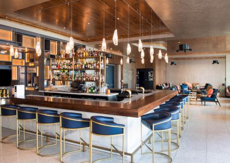 A modern bar with seating, a stocked liquor shelf, stylish pendant lights, and a cozy lounge area in the background.