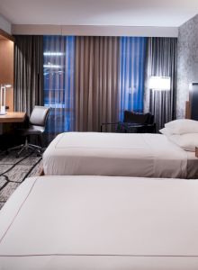 The image shows a modern hotel room with two beds, a desk with a chair, a large TV, and stylish decor including patterned walls and curtains.