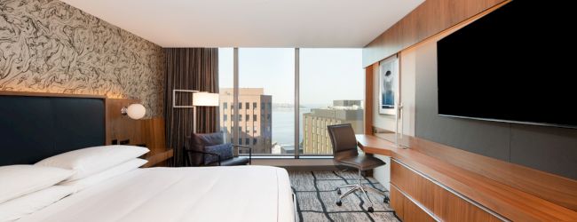 A modern hotel room with a bed, large TV, desk, and chair. The window shows buildings and a water view in the background.