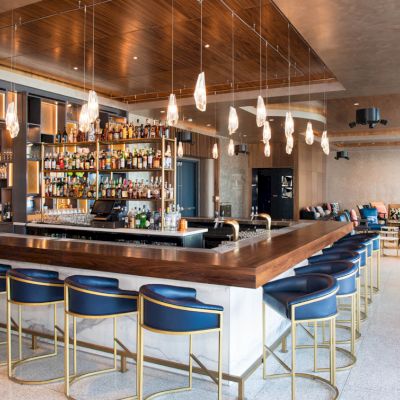 The image shows a modern bar with a sleek counter, blue stools, hanging lights, and a well-stocked liquor shelf. There are seating areas.