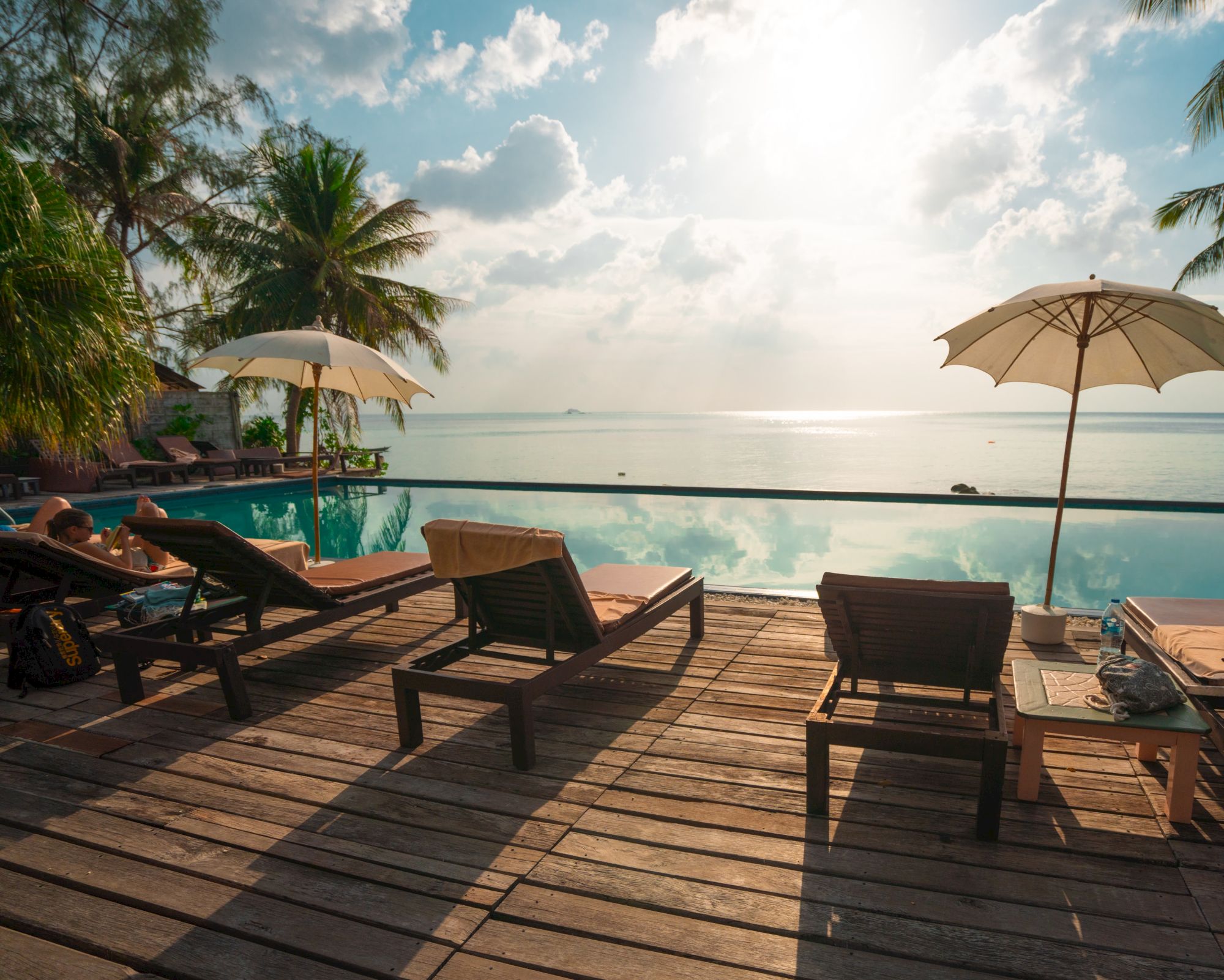 A serene beach scene with lounge chairs, umbrellas, and an infinity pool overlooking the ocean under a bright sky.