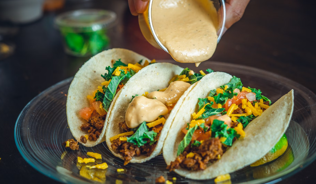 Three tacos with toppings like cheese and greens are being drizzled with a creamy sauce from a cup.