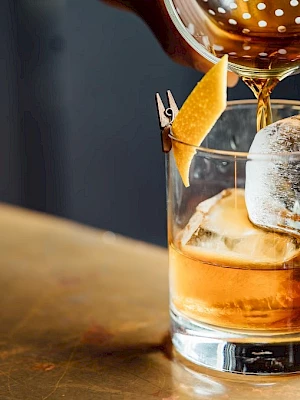 A drink being poured into a glass with ice and a citrus garnish on a metal surface.