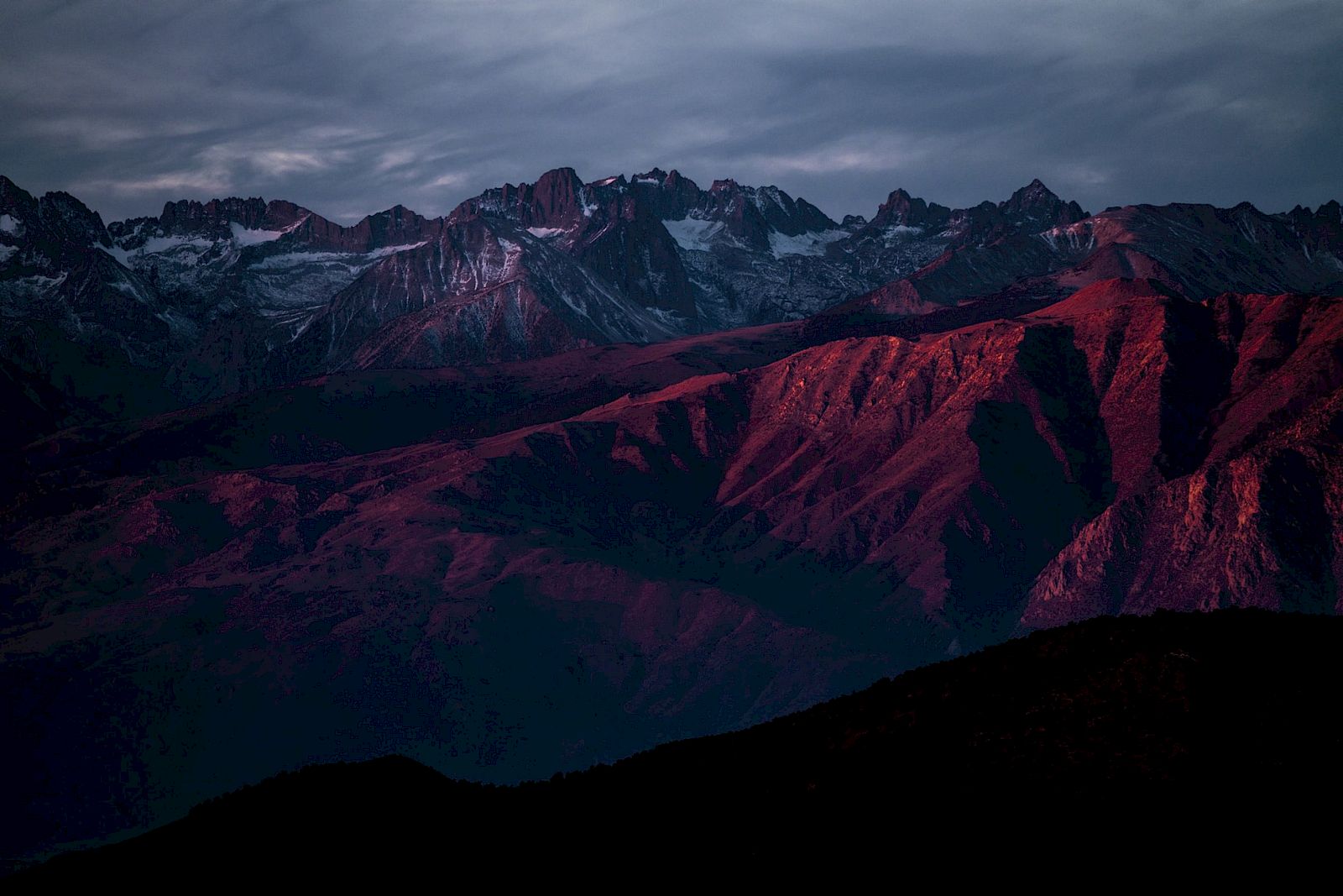 A mountain range with rugged peaks is bathed in red light, under a cloudy sky, creating a dramatic and atmospheric scene.