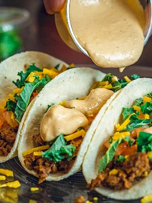 A hand is pouring sauce over tacos filled with ground meat, cheese, tomatoes, and greens, all on a glass plate.