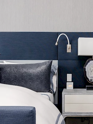 A modern bedroom features a blue upholstered headboard, decorative pillows, a nightstand with a stylish lamp, and wall lighting.