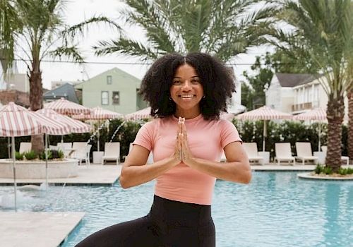 A person is practicing yoga by a pool, standing in a tree pose with palms together, surrounded by palm trees and lounge chairs.