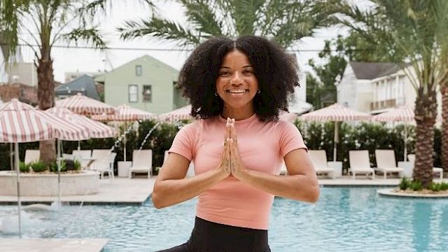 A person is practicing yoga by a pool, standing in a tree pose with palms together, surrounded by palm trees and lounge chairs.