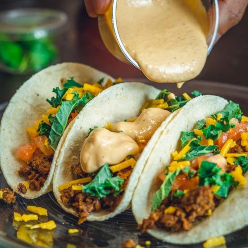 Three tacos filled with ground meat, cheese, lettuce, and tomato, being topped with creamy sauce on a glass plate.