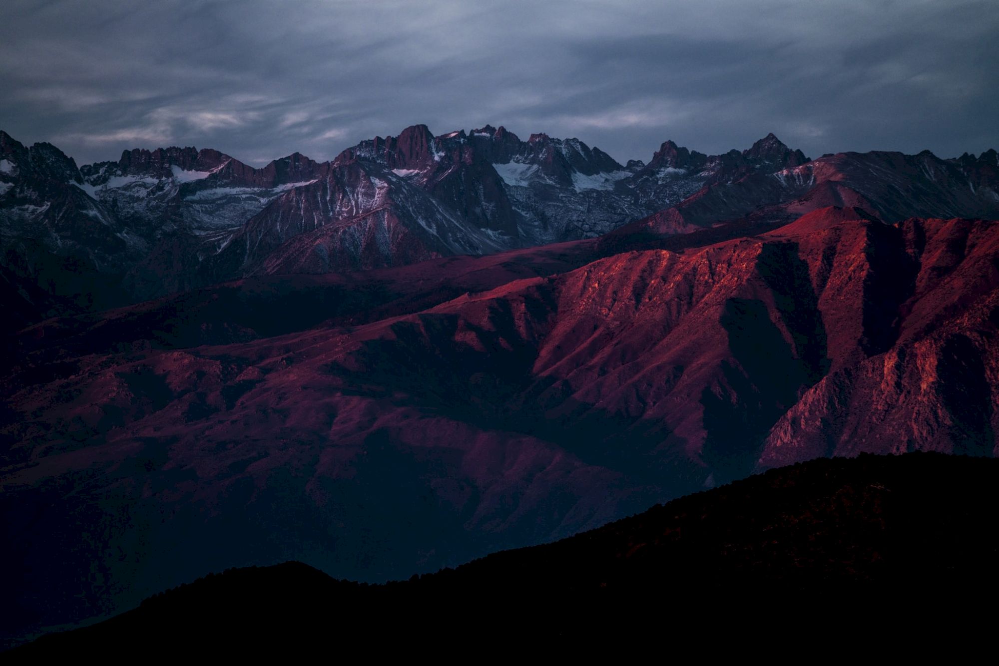 A mountain range at dusk with peaks illuminated by red sunlight and a cloudy sky above.