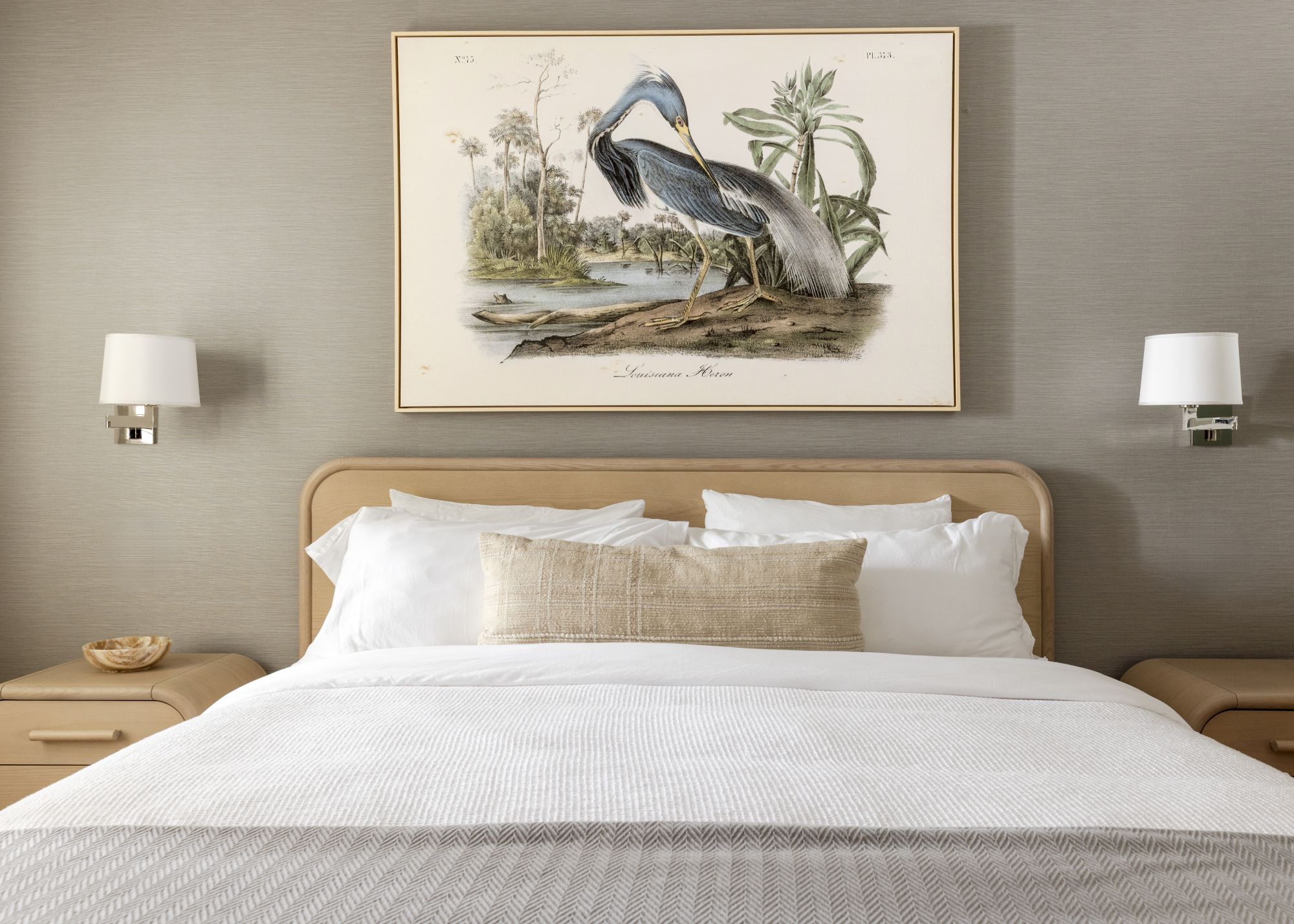 A neatly made bed with white linens and a beige pillow, flanked by two wooden nightstands. A bird artwork hangs above the bed.