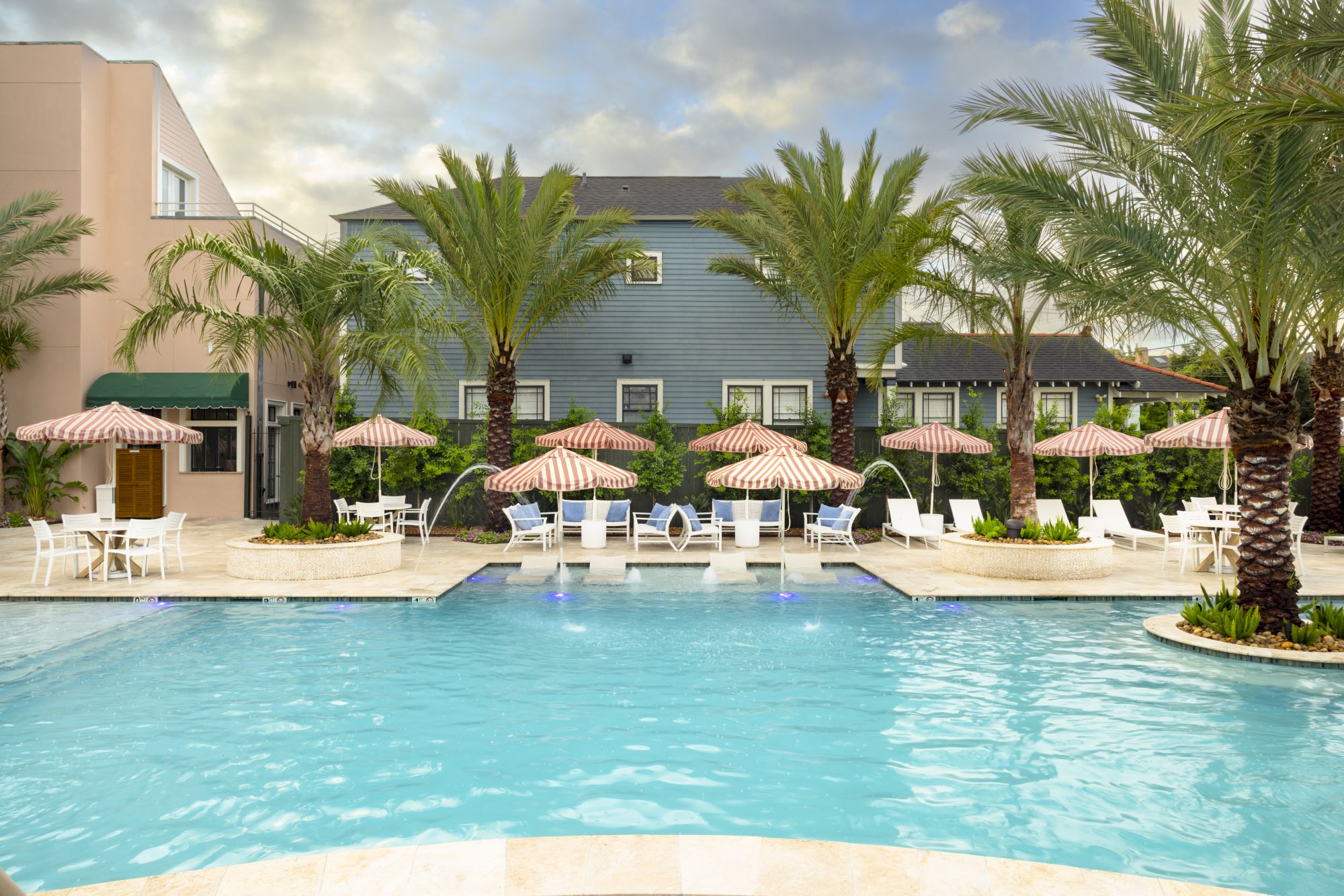 A serene poolside scene with palm trees, sun loungers, striped umbrellas, and a building in the background under a cloudy sky.
