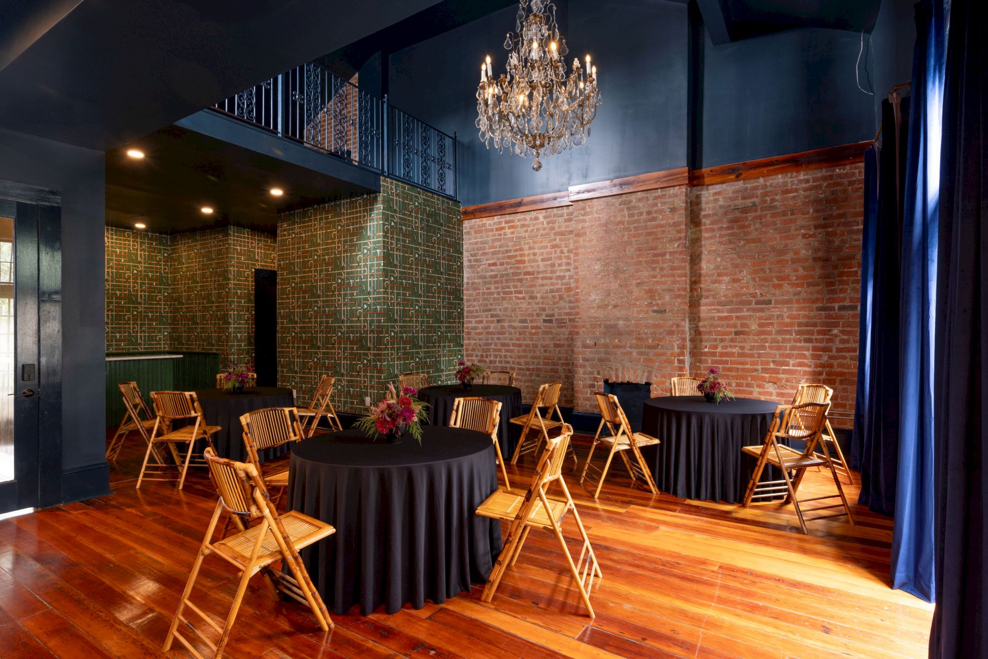 An elegant event room with a crystal chandelier, round tables, bamboo chairs, a red brick wall, and patterned panels.