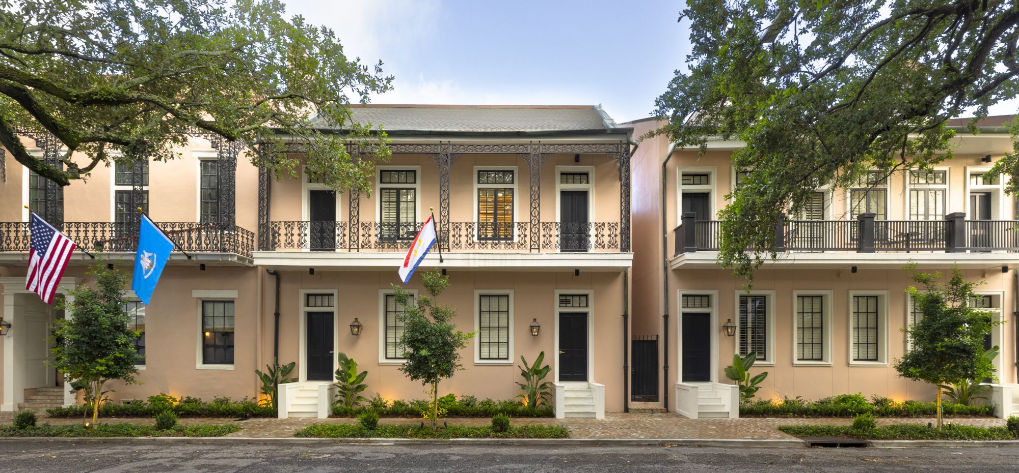 Row of peach-colored buildings with black wrought iron balconies, flags, and small trees in front; a peaceful street scene.