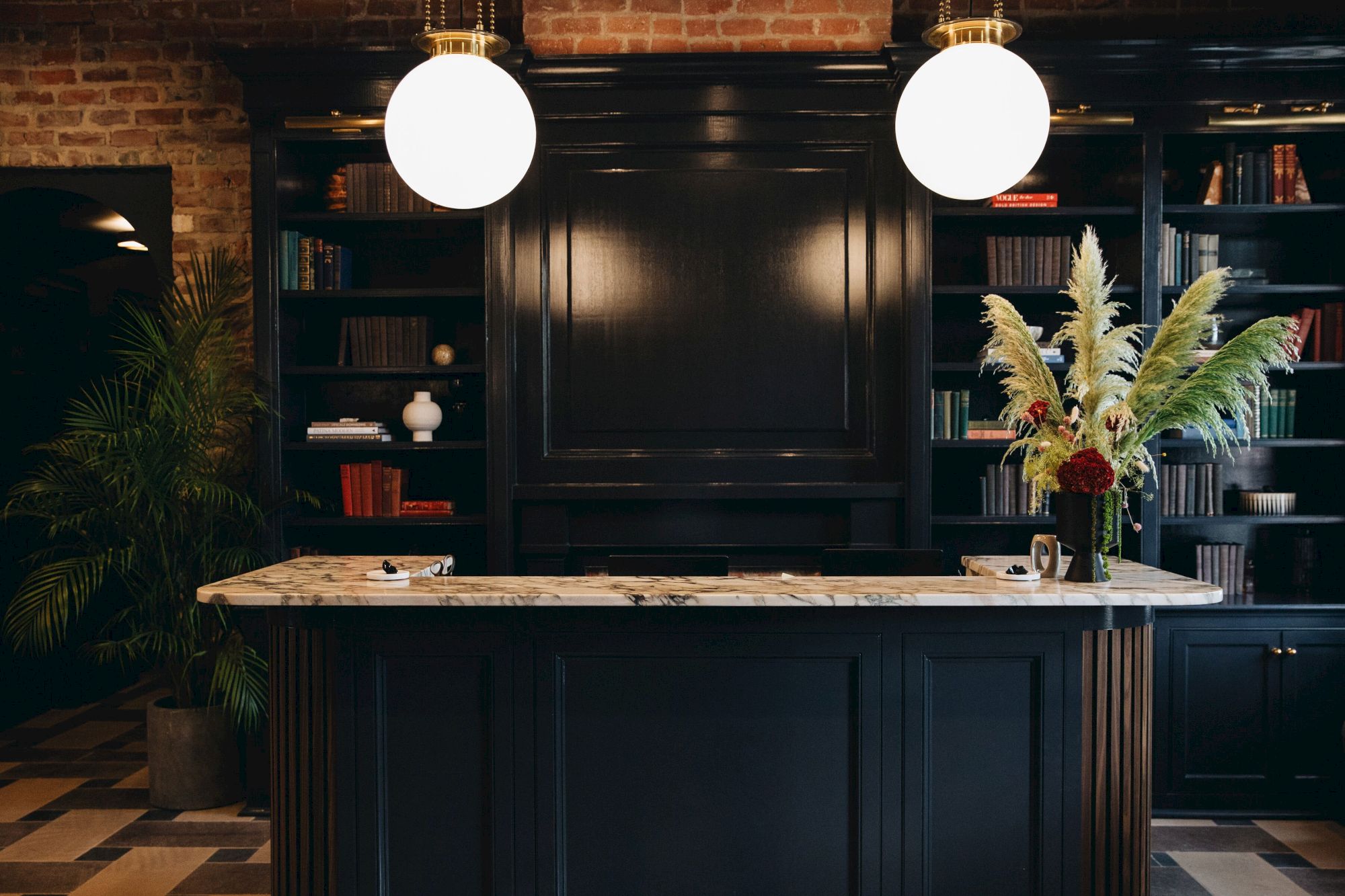 A stylish reception area with a marble counter, globe lights, and dark shelves filled with books and decorative plants.