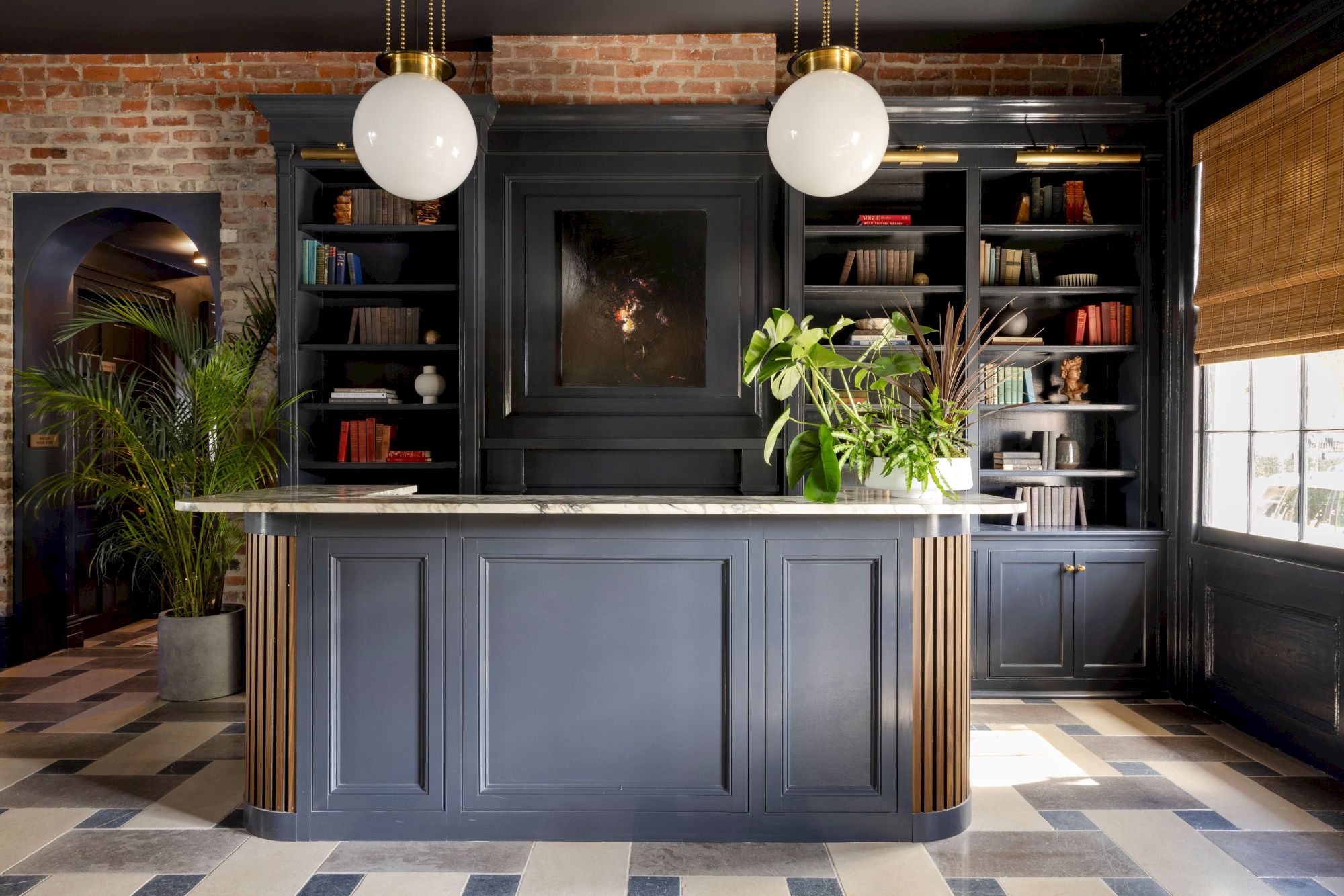The image shows a stylish room with a navy counter, shelves with books, plants, and two round lights. Brick walls add a rustic touch.