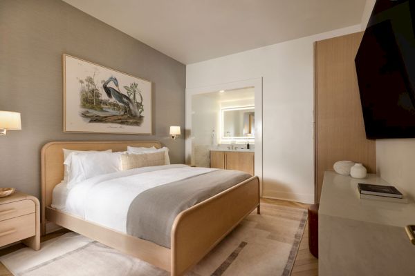 A cozy bedroom with a large bed, soft lighting, wall art of a pelican, and a TV on the wall. A glimpse of a bathroom is visible.