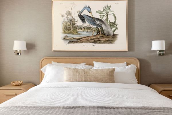 A cozy bedroom with a neatly made bed, flanked by two nightstands and wall lamps, featuring a framed bird illustration above.