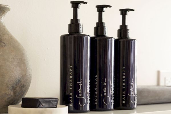 Three dark pump bottles labeled "Botanical" and "Hair Therapy" are on a shelf with a small stone dish and a vase.