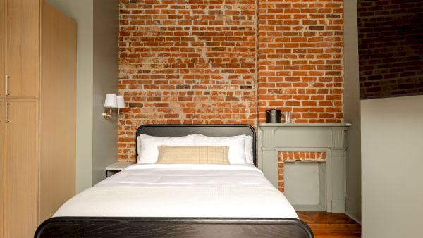 A cozy bedroom with a brick wall, bed, beige wardrobe, and wall lamp, creating a warm and inviting atmosphere.