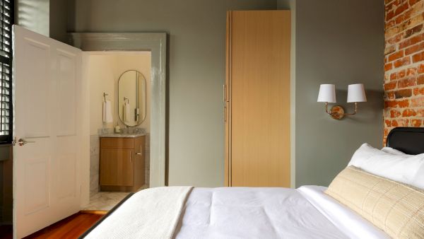 A cozy bedroom with a bed, wooden floor, exposed brick wall, and an open door leading to a bathroom with a sink and mirror.