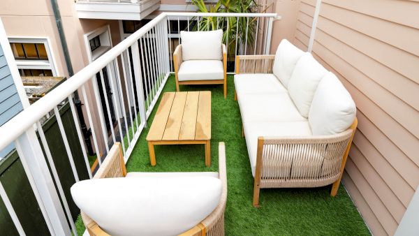 A cozy balcony with white cushioned patio furniture on artificial grass and wooden accents, flanked by railings and exterior walls.
