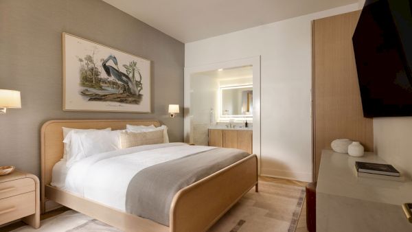 A modern bedroom with a painting, a large bed, and soft lighting, leads to an open bathroom inside.