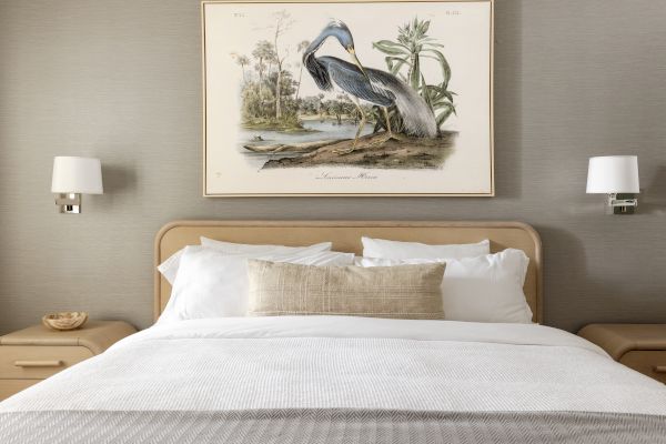 A neatly made bed with white linens is in front of a wall featuring a framed bird illustration and two wall-mounted lamps.