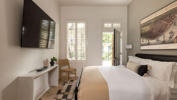 A modern bedroom with a large bed, mounted TV, console table, art on the wall, chair, and window shutters.