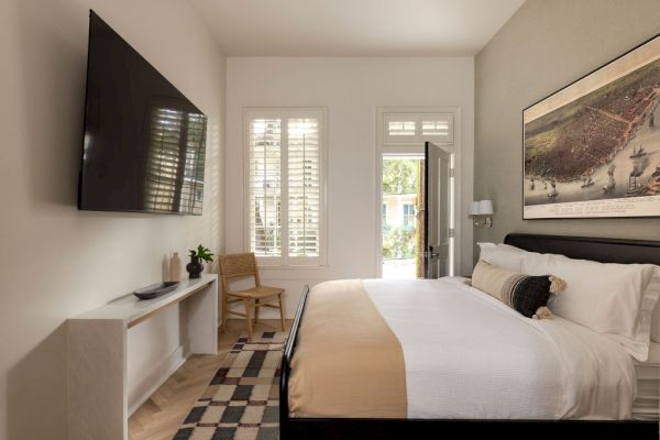 A modern bedroom with a large bed, mounted TV, console table, art on the wall, chair, and window shutters.