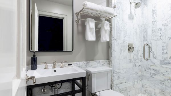 A modern bathroom with a sink, mirror, toilet, and glass shower enclosure featuring marble tiles and a towel rack above the toilet.
