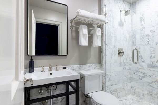 A modern bathroom with a sink, mirror, toilet, and glass shower enclosure featuring marble tiles and a towel rack above the toilet.