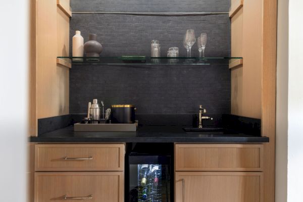 A small bar nook with wooden cabinets, a glass shelf with decor, glasses, a sink, and a mini fridge below the counter is seen in this image.