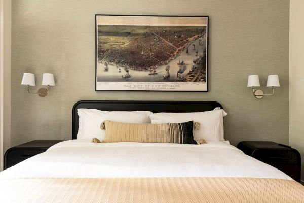 A neatly made bed with a tan blanket and decorative pillows, two wall lamps, and a framed artwork above the headboard.