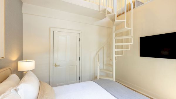 A cozy bedroom with a bed, a lamp, a wall-mounted TV, and a spiral staircase leading to a loft area.