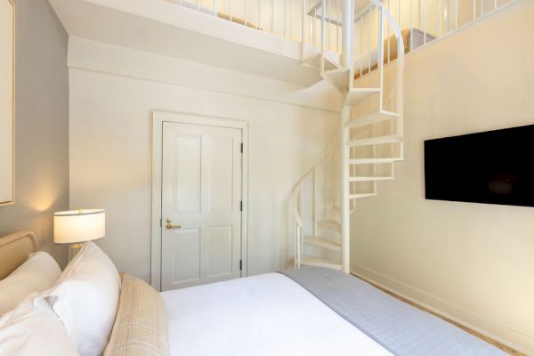 A cozy bedroom with a bed, a lamp, a wall-mounted TV, and a spiral staircase leading to a loft area.