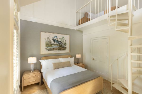 A cozy bedroom with a loft features a bed, two nightstands with lamps, a spiral staircase, and a pelican artwork on the wall.