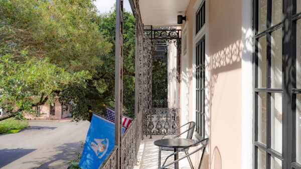 A balcony with intricate railings, a small table, and chairs overlooks a street. Two flags hang from the railing on a sunny day.