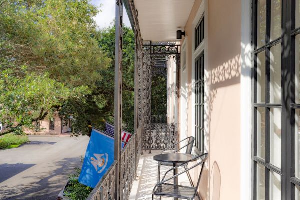 A balcony with intricate railings, a small table, and chairs overlooks a street. Two flags hang from the railing on a sunny day.