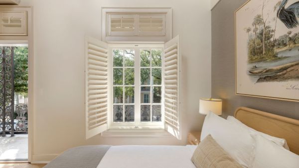 A cozy bedroom with an open window, white shutters, a bed with gray and white bedding, and a nature-themed picture on the wall.