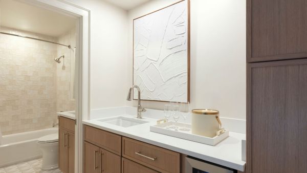 Modern kitchenette with wood cabinets, white countertop, abstract wall art, and a small fridge; a bathroom is visible in the background.